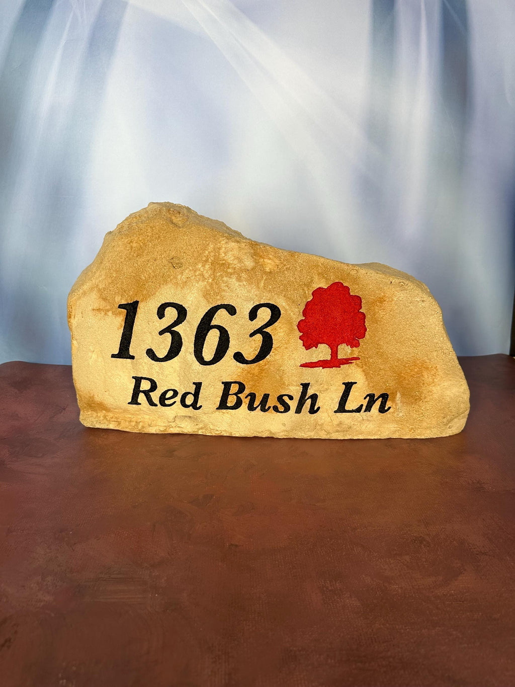 A large, tan landscape stone with an irregular, rounded shape. The left hand side of the stone is slightly taller than the right size of the stone. The stone sits upright giving a nice face to engrave upon. The stone has a rustic, handcrafted appearance with natural texture and subtle color variations. This stone has an address with street name engraved in black. There is also a tree that is painted red.