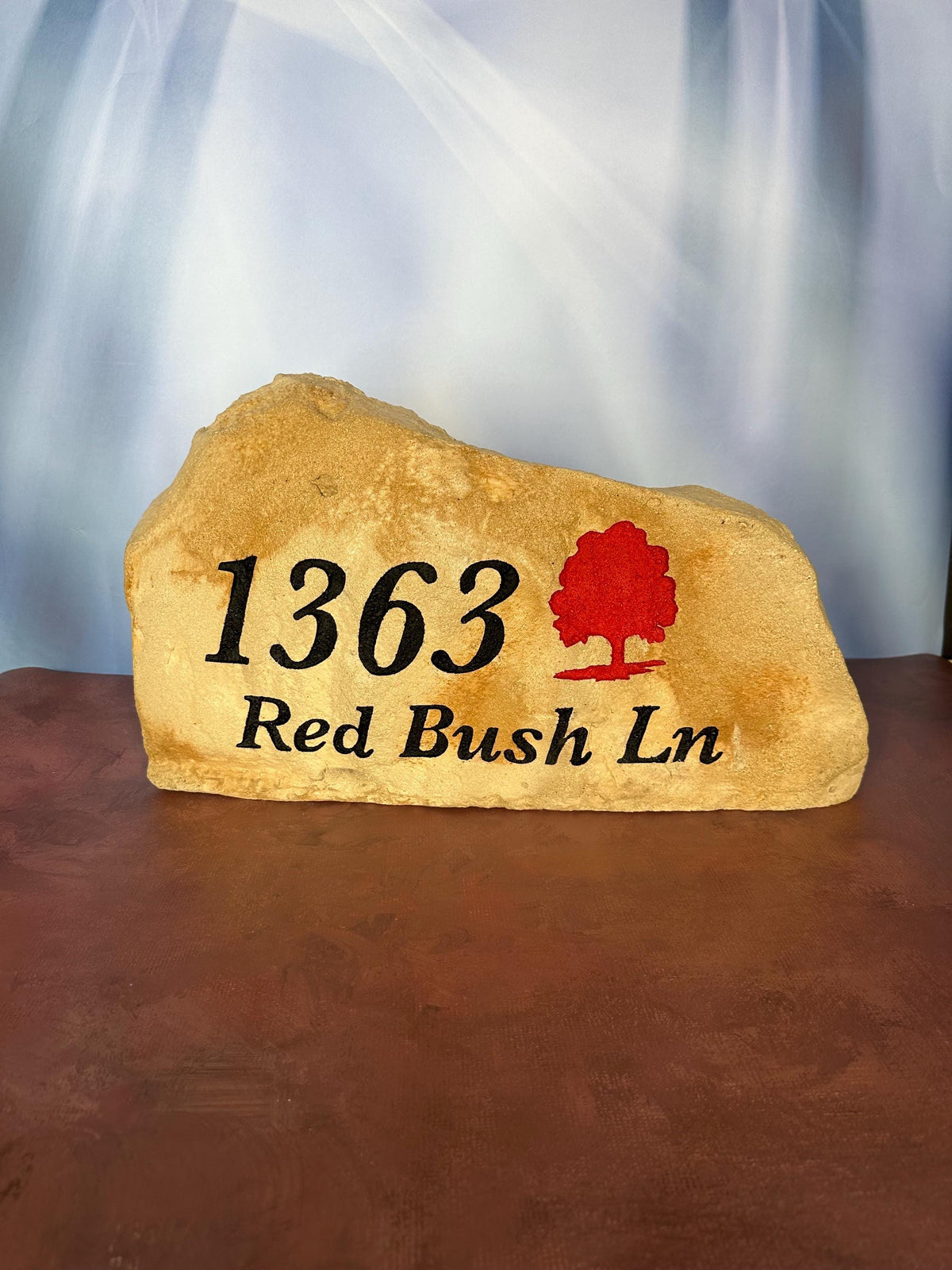 A large, tan landscape stone with an irregular, rounded shape. The left hand side of the stone is slightly taller than the right size of the stone. The stone sits upright giving a nice face to engrave upon. The stone has a rustic, handcrafted appearance with natural texture and subtle color variations. This stone has an address with street name engraved in black. There is also a tree that is painted red.