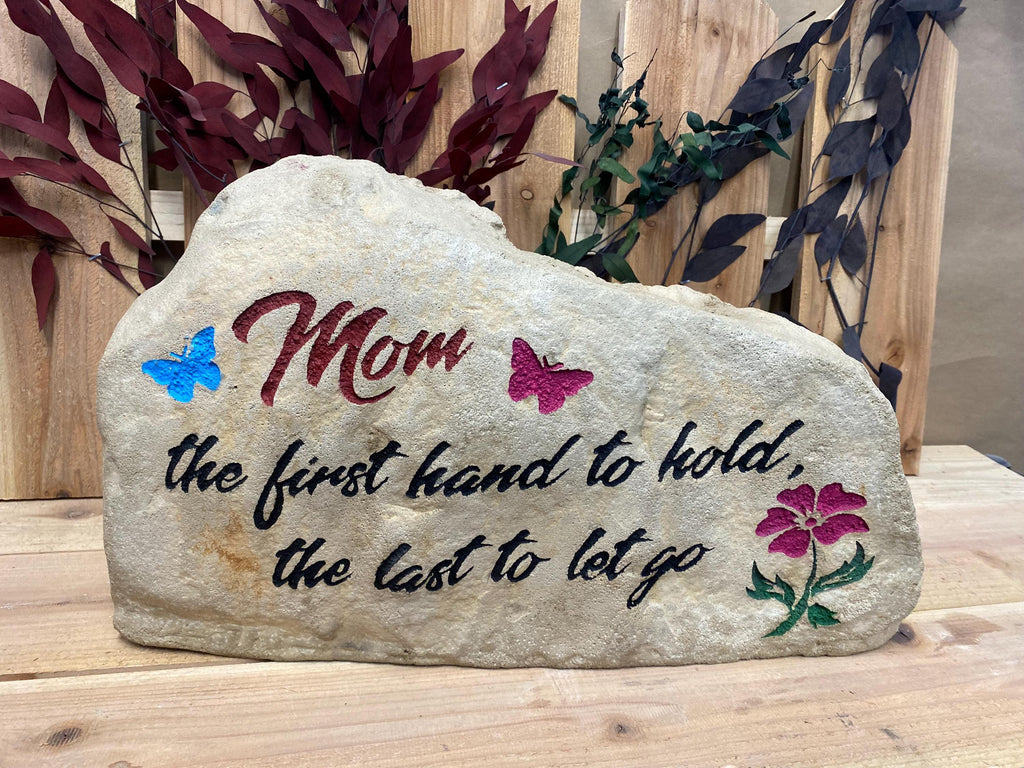 This stone has the text Mom the first hand to hold the last to let go. Mom is in a dark pink color and the rest of the text is in black. There are also two butterflies in blue and dark pink. there is a dark pink flower with a green stem.