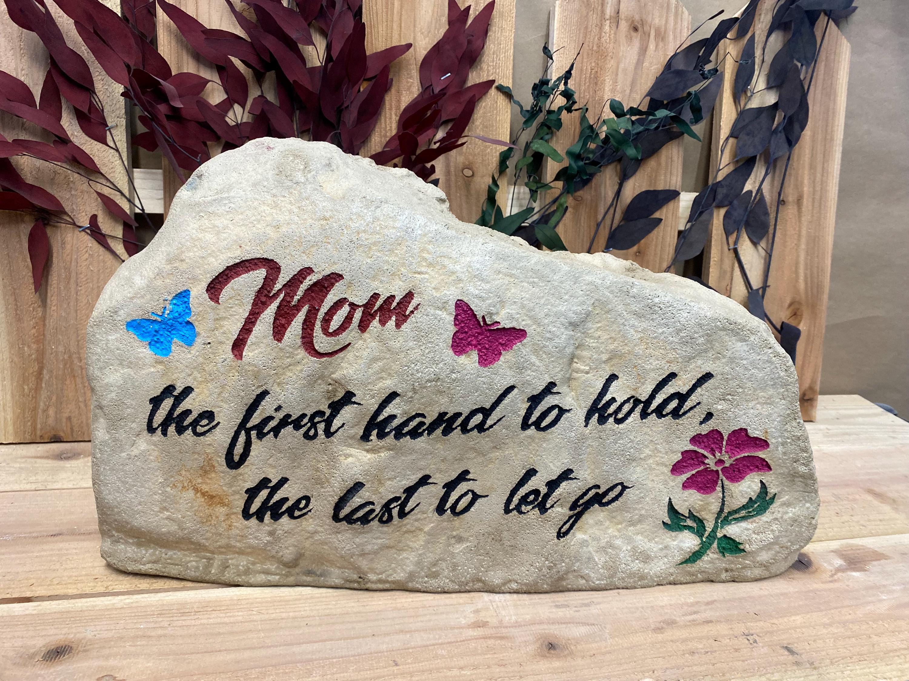 This stone has the text Mom the first hand to hold the last to let go. Mom is in a dark pink color and the rest of the text is in black. There are also two butterflies in blue and dark pink. there is a dark pink flower with a green stem.