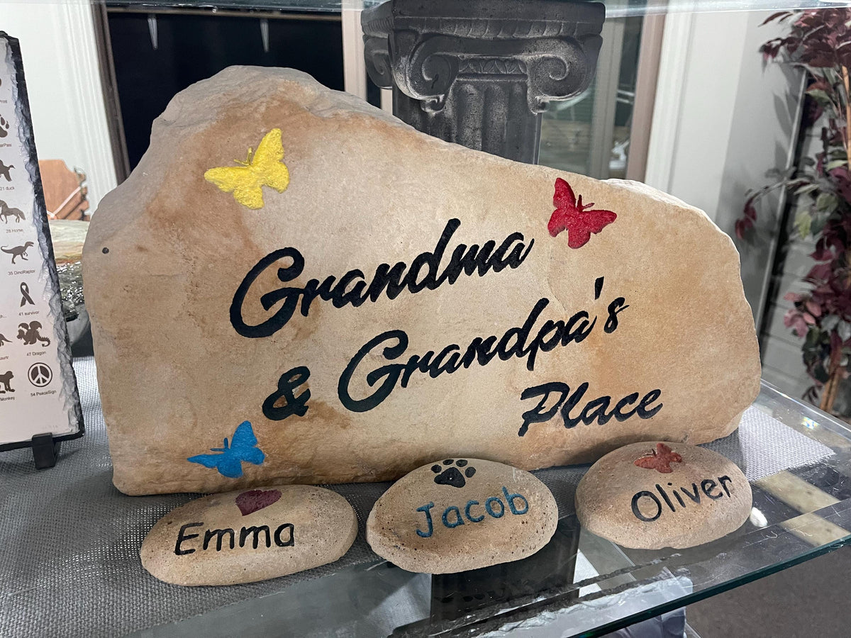 A large, tan landscape stone with an irregular, rounded shape. The left hand side of the stone is slightly taller than the right size of the stone. The stone sits upright giving a nice face to engrave upon. This stone has the text Grandma & Grandpa's place written on it with three butterflies. The text is black and the butterflies are pink, yellow, and blue.